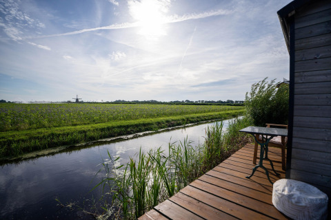 Blick von einer Holzterrasse auf einen Kanal, grüne Felder und eine Windmühle nahe der niederländischen Küste.