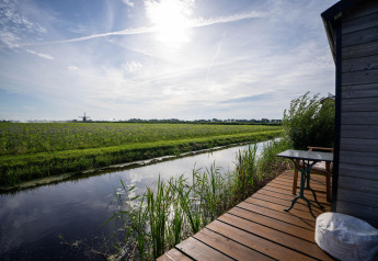 Udsigt over en kanal og grønne marker fra terrassen ved et tiny house nær kysten i Holland, klar himmel.