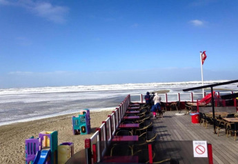 Zicht vanaf een caféterras aan de kust in Nederland met strand, zee en een speeltuin op het zand.