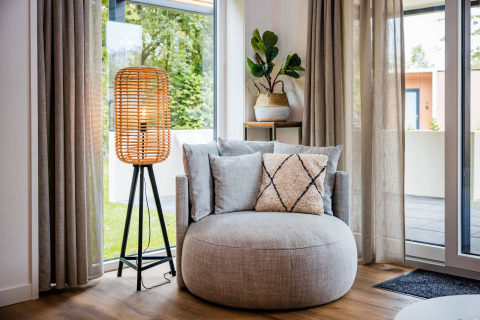 Hyggelig lodge-stue med rund lænestol, gulvlampe, grøn plante og store vinduer med gardiner.