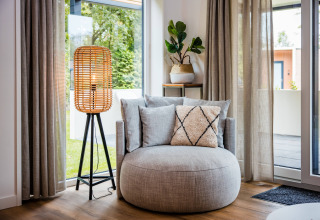 Hyggelig lodge-stue med rund lænestol, gulvlampe, grøn plante og store vinduer med gardiner.