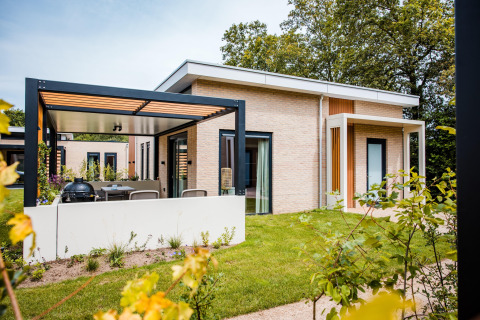 Moderne Lodge mit überdachter Terrasse, Garten und großen Fenstern, umgeben von grüner Natur.