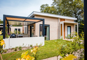 Moderne Lodge mit überdachter Terrasse, Garten und großen Fenstern, umgeben von grüner Natur.