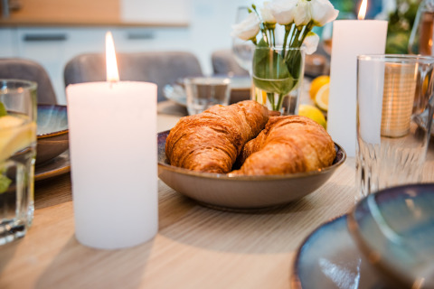 Tavola della colazione in un lodge con croissant in una ciotola, candele accese e fiori freschi sullo sfondo.