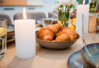 Tavola della colazione in un lodge con croissant in una ciotola, candele accese e fiori freschi sullo sfondo.