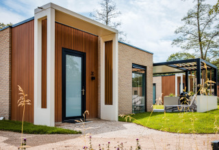Moderne lodge med træ- og stenelementer, store vinduer, terrasse og grønne omgivelser under blå himmel.
