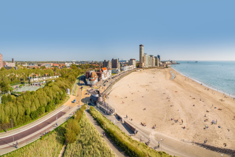 Panoramablick auf einen Sandstrand mit Menschen, Hochhäusern und Grünflächen unter klarem Himmel.