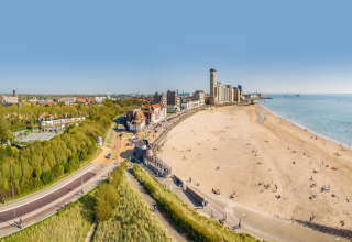 Panoramic view of a sandy beach with people, coastal buildings, and green spaces on a clear day.