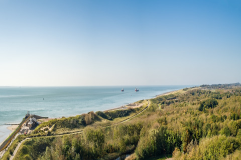 Panoramic view of a beach house, lush dunes and the coastline stretching along a calm, blue sea.
