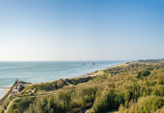 Panoramic view of a beach house, lush dunes and the coastline stretching along a calm, blue sea.
