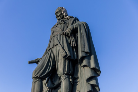 Statue of a historical figure in a cape holding a scroll, photographed against a clear blue sky.