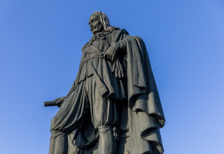 Statue of a historical figure in a cape holding a scroll, photographed against a clear blue sky.