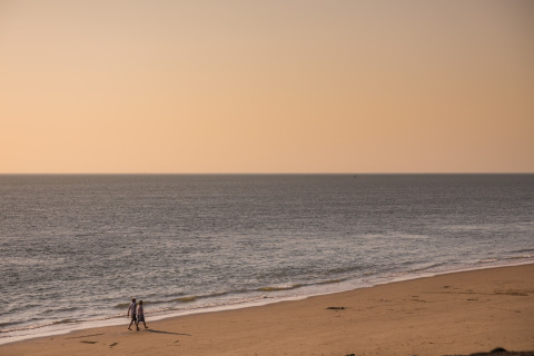 Two people walk along a quiet beach at sunset, with soft light illuminating the sea and sand.