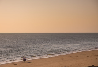 Two people walk along a quiet beach at sunset, with soft light illuminating the sea and sand.