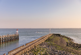 Blick auf eine lange Mole am Strand, die ins Meer führt, mit ruhigem Wasser und einem Boot im Hintergrund.