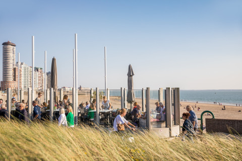 People dining and relaxing at a modern beach house terrace, grassy dunes and city buildings in view.