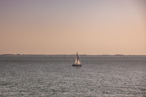 Ein Segelboot fährt ruhig auf dem Meer bei Sonnenuntergang, gesehen von einem Strandhaus aus.