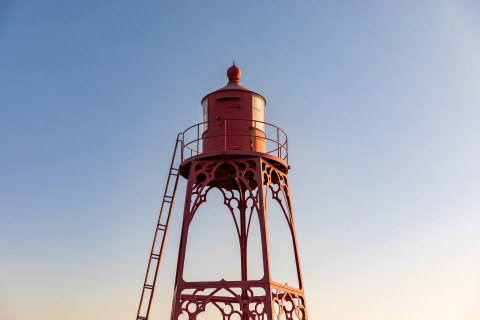Ein roter Leuchtturm aus Metall steht am Strandhaus vor klarem Himmel im warmen Abendlicht.