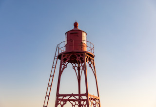 Ein roter Leuchtturm aus Metall steht am Strandhaus vor klarem Himmel im warmen Abendlicht.