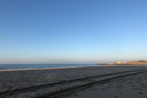 Peaceful beach at sunrise with tire tracks in the sand, calm sea, and a distant house by the shore.