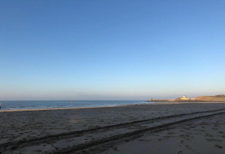 Peaceful beach at sunrise with tire tracks in the sand, calm sea, and a distant house by the shore.