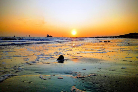 Sonnenuntergang am Strand vor einem Strandhaus, schäumende Wellen und ein Schiff am Horizont.
