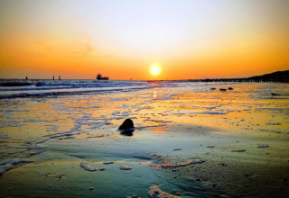 Sonnenuntergang am Strand vor einem Strandhaus, schäumende Wellen und ein Schiff am Horizont.