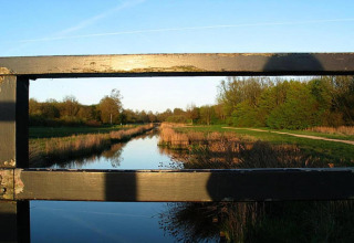 Blick von einer Holzbrücke auf einen ruhigen Fluss und grüne Landschaft unter klarem Himmel.