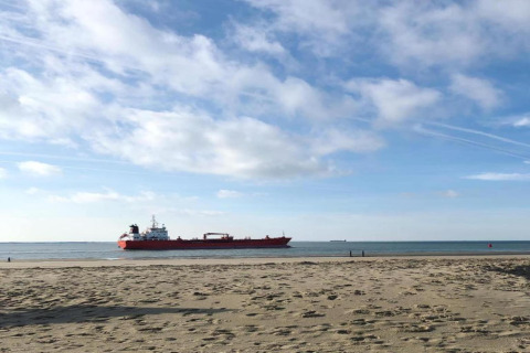Aussicht von einem Strandhaus auf ein großes rotes Frachtschiff, das bei blauem Himmel auf dem Meer fährt.
