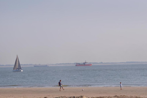 Two people play football on a sandy beach, with sailboats and a cargo ship in the calm sea background.