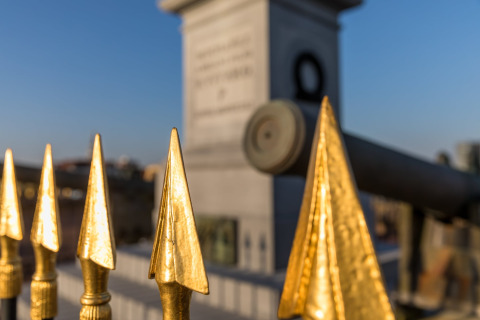 Golden fence spikes in the foreground with a cannon and stone monument blurred in the background.