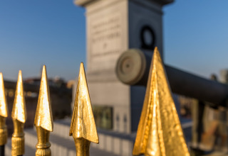 Golden fence spikes in the foreground with a cannon and stone monument blurred in the background.