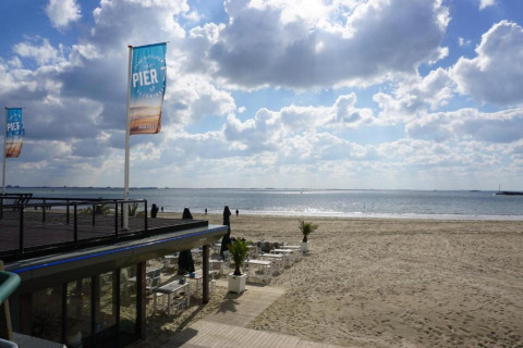 Beach house with outdoor seating and Pier 7 flag, facing sandy beach and sea under a partly cloudy sky.