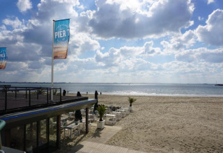Strandhaus mit Außensitzplätzen und Pier 7-Flagge, Blick auf Sandstrand und Meer unter bewölktem Himmel.