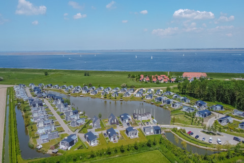 Luftfoto af moderne lodges ved en sø og havet, omkranset af natur, med blå himmel og både i baggrunden.