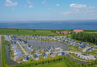 Luftfoto af moderne lodges ved en sø og havet, omkranset af natur, med blå himmel og både i baggrunden.