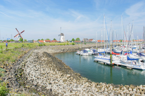 Både ligger fortøjet i en havn med vindmøller og farverige tage i baggrunden under en blå himmel.