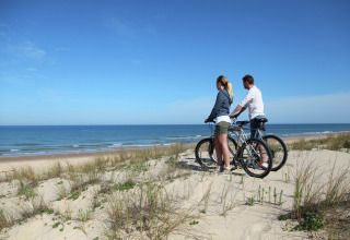 To personer på cykler nyder udsigten over havet fra sandklitterne ved en lodge under blå himmel.