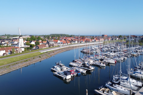 Både i en marina ved siden af en landsby med en vindmølle og røde tage under en klar blå himmel.