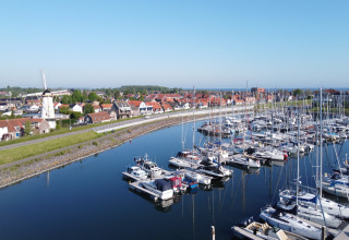 Både i en marina ved siden af en landsby med en vindmølle og røde tage under en klar blå himmel.