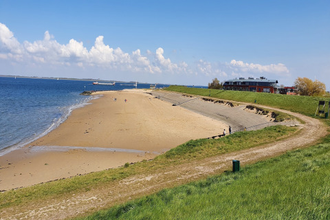 En rolig strand med en lodge i baggrunden, grønne skråninger og få mennesker under en blå himmel.