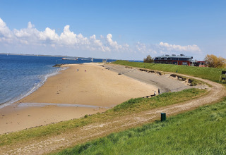 En rolig strand med en lodge i baggrunden, grønne skråninger og få mennesker under en blå himmel.