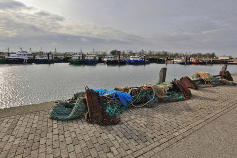 Både ligger fortøjet i en havn med fiskegarn og udstyr på kajen under en overskyet himmel.