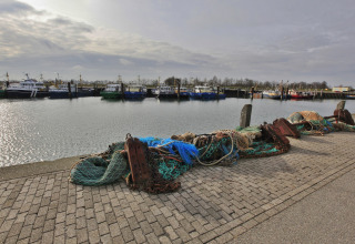 Både ligger fortøjet i en havn med fiskegarn og udstyr på kajen under en overskyet himmel.