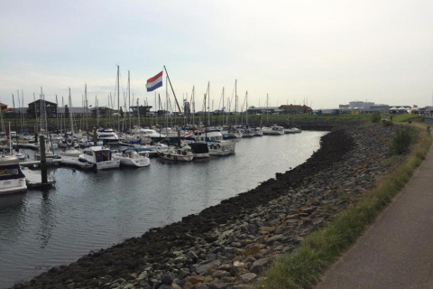 Både fortøjet i en marina ved en lodge med en stor hollandsk flag og promenade til højre.