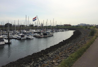 Både fortøjet i en marina ved en lodge med en stor hollandsk flag og promenade til højre.
