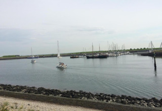 Både sejler ved en marina med flere fortøjede både, stenmole og grøn baggrund ved en lodge.
