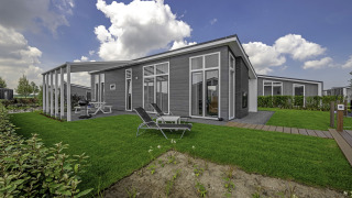 Moderne lodge ved Water Resort Oosterschelde 34 med store vinduer, terrasse og grøn have under blå himmel.
