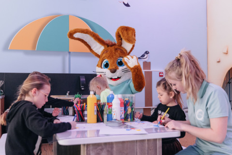 Børn og voksen laver kunsthåndværk med en kaninmaskot ved et bord på et glampingophold for familier.