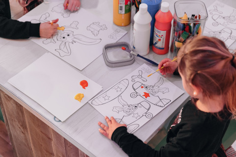 Children painting coloring sheets at a table filled with paints and art supplies in a glamping setting.